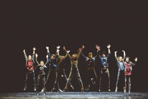 Group of young performers on the stage standing in the line, jumping with raised hands. Dark tones.
